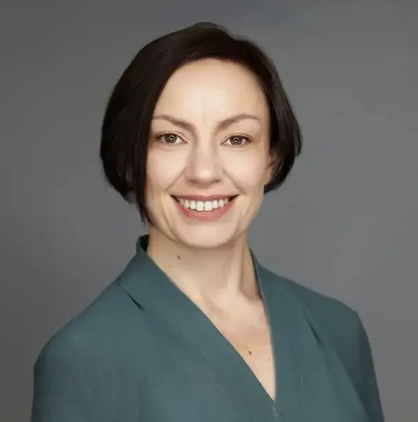 Porträt einer freundlich lächelnden Frau mit dunklem, kurzem Haar vor grauem Hintergrund.