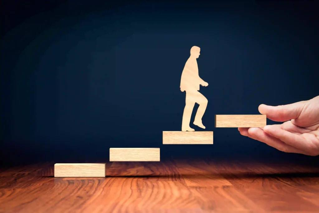 Holzfigur auf einer Treppe aus Holzklötzen, während eine Hand die nächste Stufe anbietet – als Symbol für individuelles Coaching und persönliche Entwicklung.