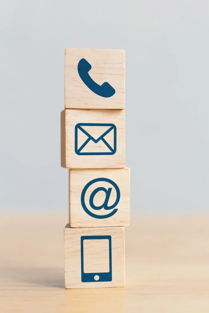 Vier übereinandergestapelte Holzklötze mit Symbolen für Telefon, E-Mail, @-Zeichen und Smartphone als Zeichen für verschiedene Kontaktmöglichkeiten.