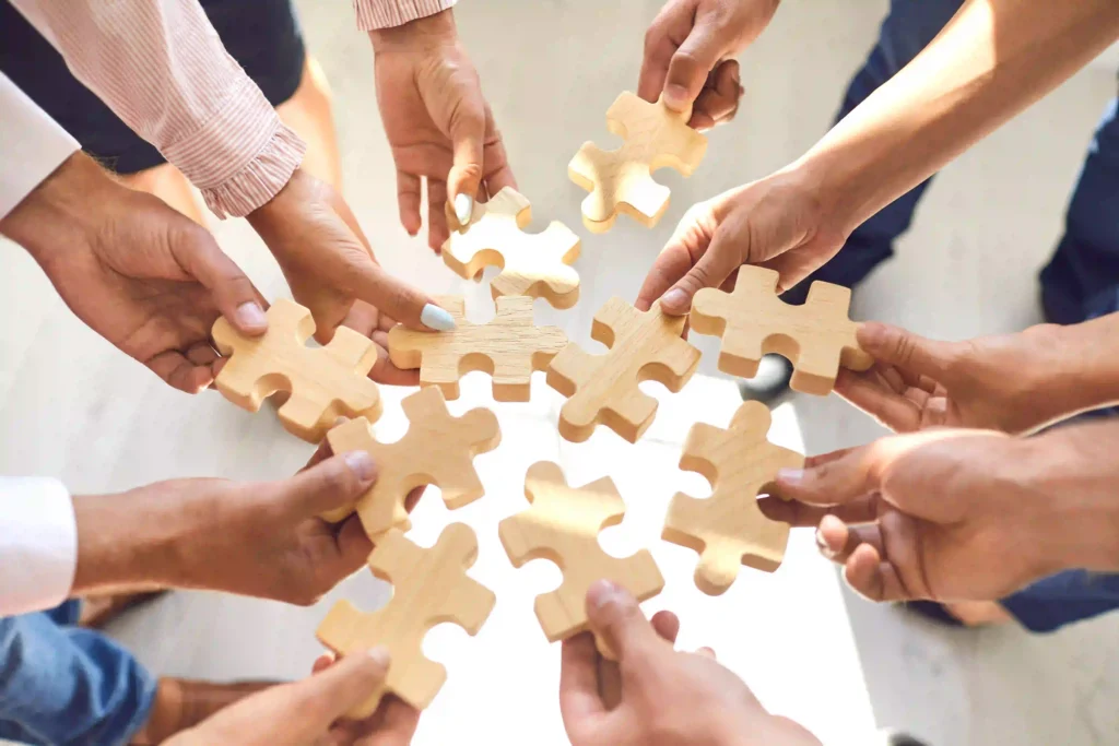 Mehrere Hände halten helle Puzzleteile zusammen und symbolisieren Teamarbeit, Zusammenarbeit und das Zusammenfügen unterschiedlicher Perspektiven.